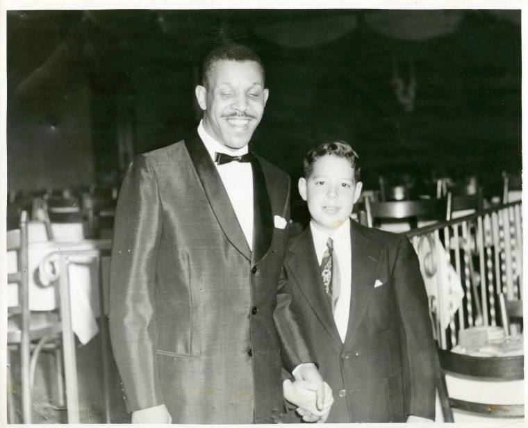 Black and white photograph of Al Hibbler standing next to a young caucasian boy holding hands. 